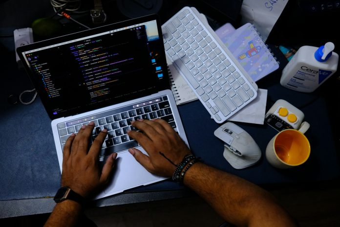 Stock | Tipkovnica i kod Ruke tipkaju na prijenosnom računalu s programskim kodom na zaslonu
